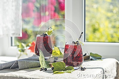 Two glass of cold ice black currant juice with ripe berries and green leaves on table in sunny room near window with garden Stock Photo