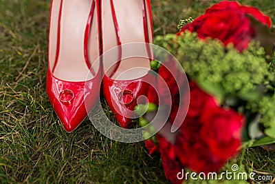 Two golden wedding rings lie on red fashion female shoes on green grass. Wedding Stock Photo