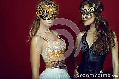 Two gorgeous young women in golden and bronze masks standing on dark red background Stock Photo