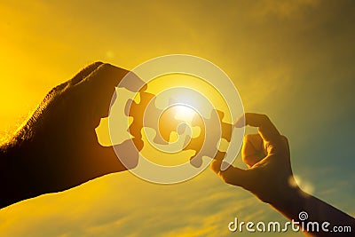 Two hands connect two pieces of the puzzle against the sky and glare of the sun at sunset. Stock Photo