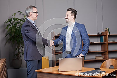 Two happy cherrful businessman in blue suits smiling and making handshake in office by the laptop, business succes concept Stock Photo