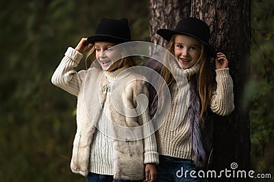 Two happy girls are equally dressed: in fur vests and hats in the forest. Little girlfriends in park. Stock Photo