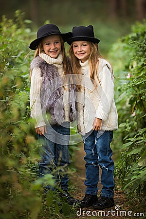 Two happy girls are equally dressed: in fur vests and hats in the forest. Little girlfriends in park. Stock Photo