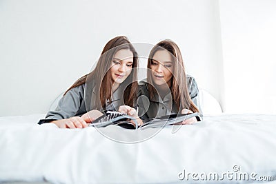 Two happy sisters twins lying on bed and reading magazine Stock Photo