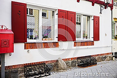 Two Italian windows on the white wall facade with open red color classic shutters and flowers on the windowsill. Stock Photo