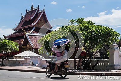 Two Laotian girls ride a scooter in front of the Wat Mai Suwannaphumaham temple in Luang Prabang, Laos, holding an umbrella Editorial Stock Photo