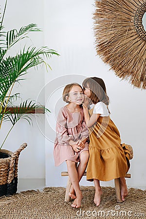 Couple of little girls of the same age sitting on a bench, whispering in the ear Stock Photo