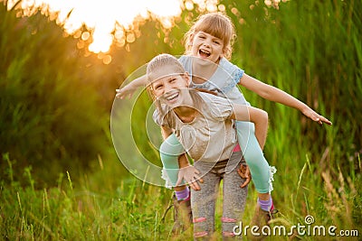 Two little funny sister playing outside on sunset. Little girl climbed up on back of cute sister and fly Stock Photo