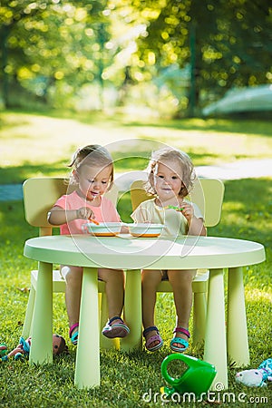 Two little girls sitting at a table and eating together against green lawn Stock Photo