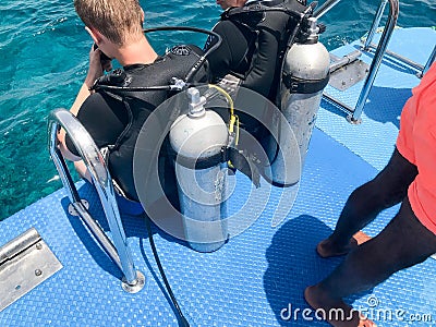 Two male divers in black diving waterproof suits with shiny metal aluminum canisters are preparing to dive from the boat to the bl Editorial Stock Photo