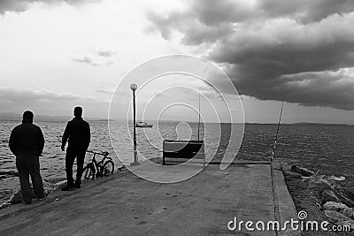 Two men are fishing on concrete pier with fishing rods. There is a bench at the pier. Photo black and white shot and cloudy sky Editorial Stock Photo