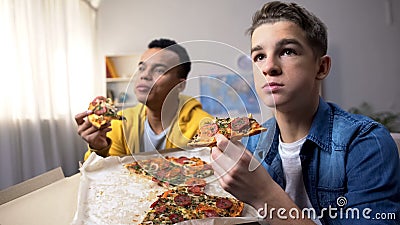 Two multiethnic male teenagers eating pizza and watching TV show, leisure time Stock Photo