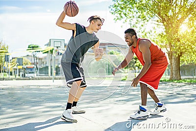 Two multiracial cheerful friends playing basketball outdoor in city urban contest with back light - Young people having fun doing Stock Photo