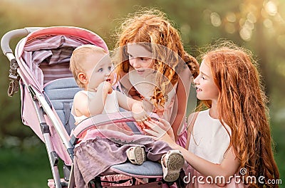 Two older redhead sisters are walking in the park on a sunny summer day with stroller with their younger sister. The girls try to Stock Photo