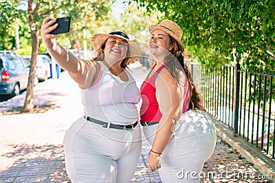 Two plus size overweight sisters twins women smiling taking a selfie picture with the phone outdoors on a sunny day Stock Photo