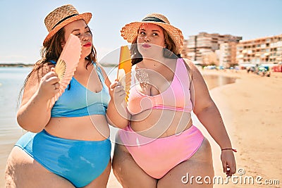 Two plus size overweight sisters twins women using handfan at beach on hoy day of summer Stock Photo