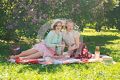 Two pretty girls friends sitting on the red blanket on the green grass and have summer picnic. happy woman having rest and fun on Stock Photo