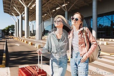 Two pretty smiling girls in sunglasses joyfully looking aside with red suitcase and backpack near airport Stock Photo