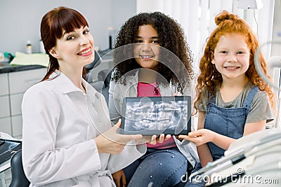 Two pretty smiling multiethnic teen girls, sitting on chair and looking at camera, while holding x-ray scan image of Stock Photo