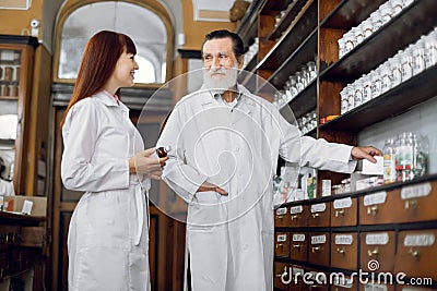 Two professional pharmacists, senior bearded man and young woman, working at ancient pharmacy. Male pharmacist holding Stock Photo