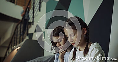 Two schoolgirls looking down sitting modern hallway. Girls watching tablet. Stock Photo
