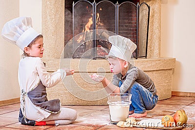 Two siblings - boy and girl - in chef`s hats near the fireplace sitting on the kitchen floor soiled with flour, playing with food Stock Photo