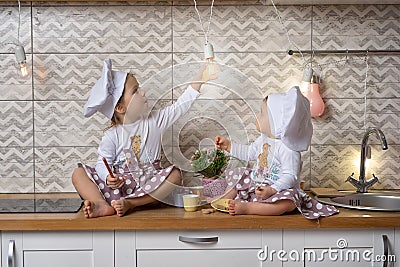 Two sisters in a cook hats sit on the kitchen table and drink tea with cookies Stock Photo
