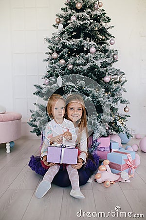 Two sisters at home with Christmas tree and presents. Happy children girls with Christmas gift boxes and decorations. Stock Photo