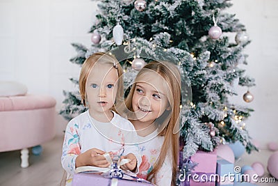 Two sisters at home with Christmas tree and presents. Happy children girls with Christmas gift boxes and decorations. Stock Photo