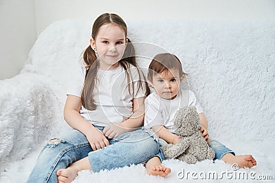 Two children, sisters sit on a white sofa in white t-shirts and blue jeans. Soft plush bear Stock Photo
