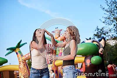 Two sisters, standing is front of attraction in theme park, holding popcorn in paper box, smiling, having fun. Pretty girls. Stock Photo