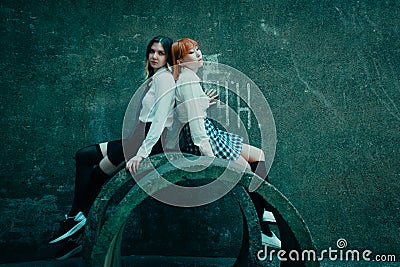 The two students in uniform sitting on a concrete ring against a gray building wall. The backdrop of a industrial city highlights Stock Photo