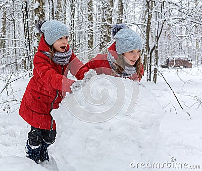 Two twin girls in red jackets rolling huge snowball to make snowman Stock Photo