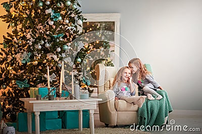 Two twin sisters sitting in a turquoise chair in the hall for the New Year. A sister whispers a secret in her ear in a decorat Stock Photo