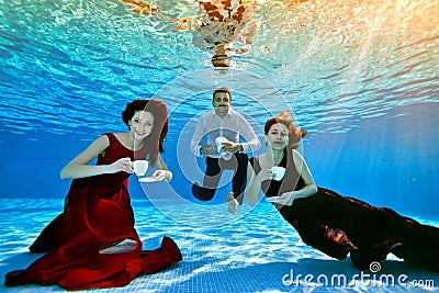 Two unusual cute girls and a guy pose and play underwater at the bottom of the pool with white cups in their hands, in Stock Photo