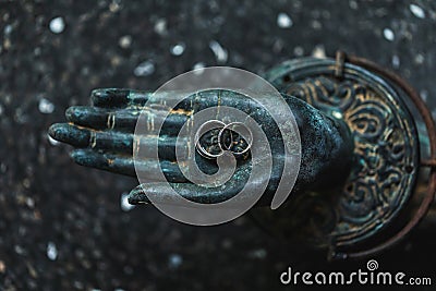 Two wedding rings on metal Buddha hand dark background. Spiritual love concept. Stock Photo