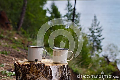 Two white metal aluminum enameled mugs with hot tea with steam on a felled wood table on the shore of Lake Baikal in a camp. Stock Photo