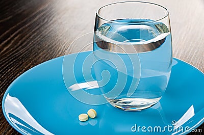 Two white tablets, glass of water in plate on wooden table Stock Photo