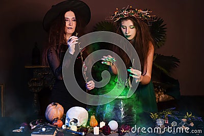Two witches with disheveled hair brew a magic potion in a black cauldron, standing in a dark room. Stock Photo