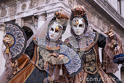 Two women in masks and costume, with decorated fans, standing in front of the arches at St Marks Square during the Venice Carnival Editorial Stock Photo