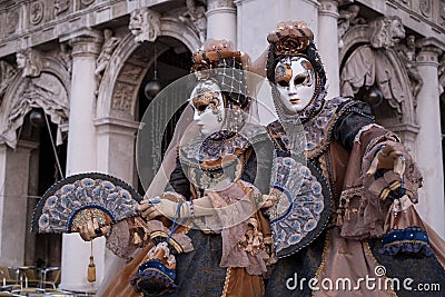 Masked women in costume, with decorated fans, standing in front of the arches at St Marks Square during the Venice Carnival Editorial Stock Photo