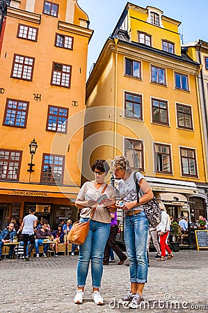 Two women tourists checking the guide on cozy medieval street with walking people, yellow buildings facades in Gamla Stan, Old Editorial Stock Photo