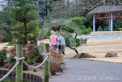 Two women walking in a Japanese garden surrounded by lush green trees and plants with yellow winter grass Editorial Stock Photo