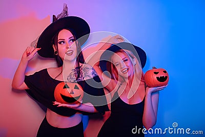 Two women witches hold pumpkins in their hands and dance. Women in neon lights are celebrating a Halloween party. Models smile and Stock Photo