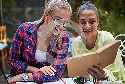 Two young adult females reading a book together, sitting in outdoor cafe, smiling, laughing Stock Photo