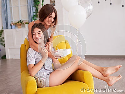 Two young brunette girls, wearing light grey cotton pajamas, sitting on yellow armchair in light apartment. Birthday celebration. Stock Photo