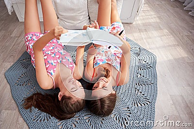 Two young brunette girls, wearing pink cotton pajamas, lying on grey carpet in light apartment, holding notebook, studying, Stock Photo