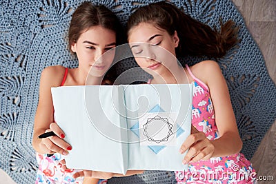 Two young brunette girls, wearing pink cotton pajamas, lying on grey carpet in light apartment, holding notebook, studying, Stock Photo