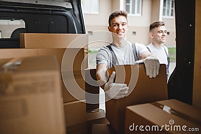 Two young handsome workers wearing uniforms are standing next to the van full of boxes. House move, mover service Stock Photo