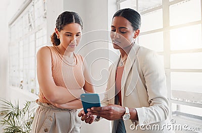 Two young happy mixed race businesswomen using a phone together at work. Hispanic businessperson talking while showing a Stock Photo